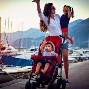 Woman holding a toddler behind a stroller with another toddler on the pier with boats in the background