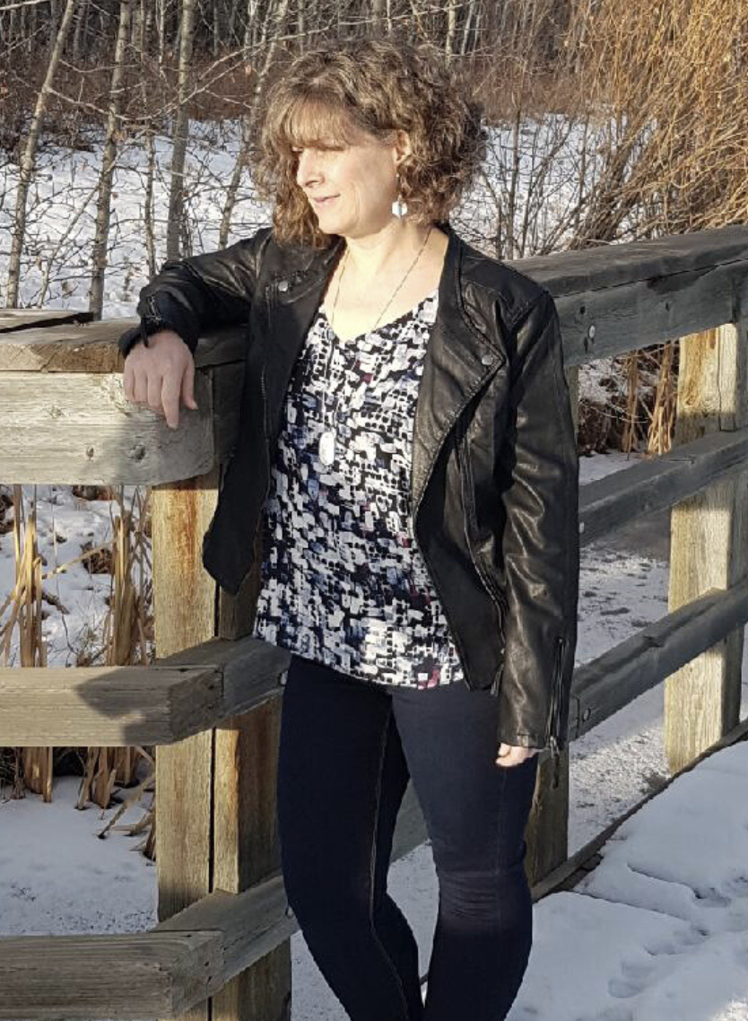 Leanne Chesser wearing blue jeans, a black leather jacket and a shirt with blue and maroon irregular shapes. She is standing with her right arm leaning on a wooden fence on a snow-covered pathway with bare trees in the background.